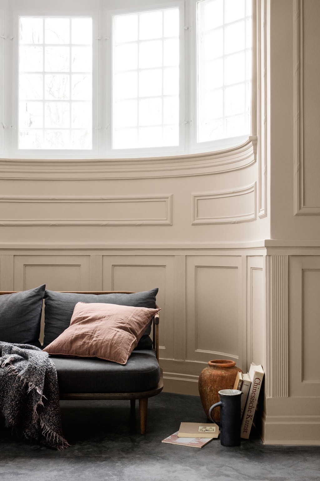 Lys stuekrok med høye sprossevindu, beige panel, mørk sofa med grå og rosa puter, vase med kopp