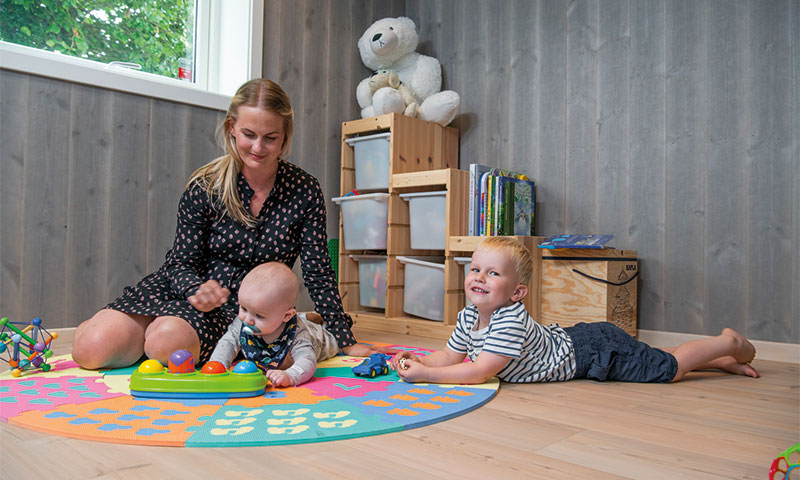 Voksen og to barn leker på fargerik sirkelmatte i lyst trelekerom med hyller og bamse