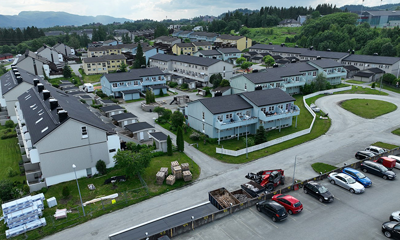 Dronebilde av boligområde med lyse rekkehus, parkerte biler, byggeutstyr og grønne åser.