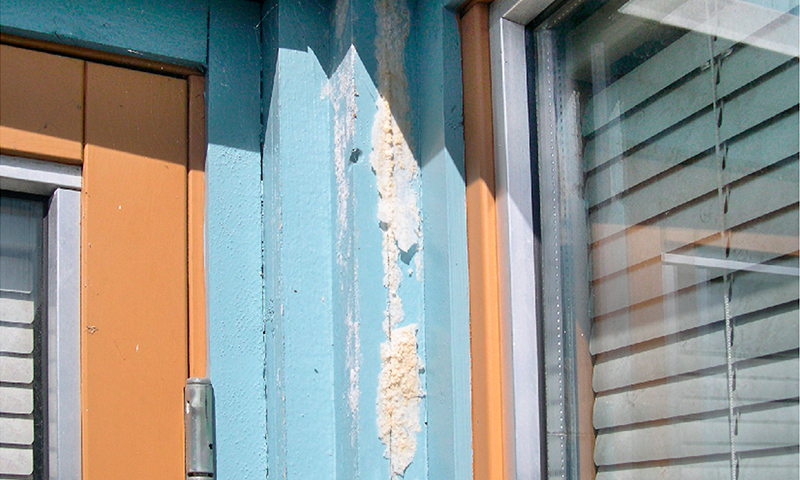 Blå søyle med avflasset maling og hvitt isolasjonsskum mellom oransje vinduskarmer.