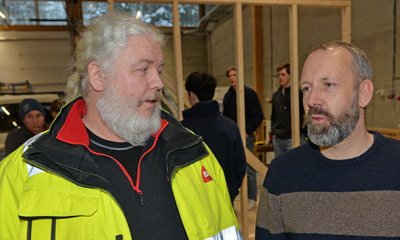 To menn i verksted foran trekonstruksjon, den ene i gul arbeidsjakke, den andre i mørk genser.