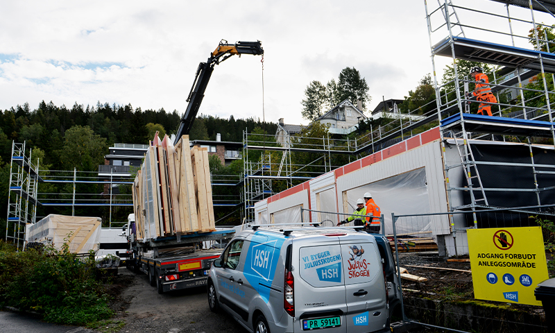 Byggeplass med stillas og hvite moduler, kran løfter trepaneler, oransjekledde arbeidere