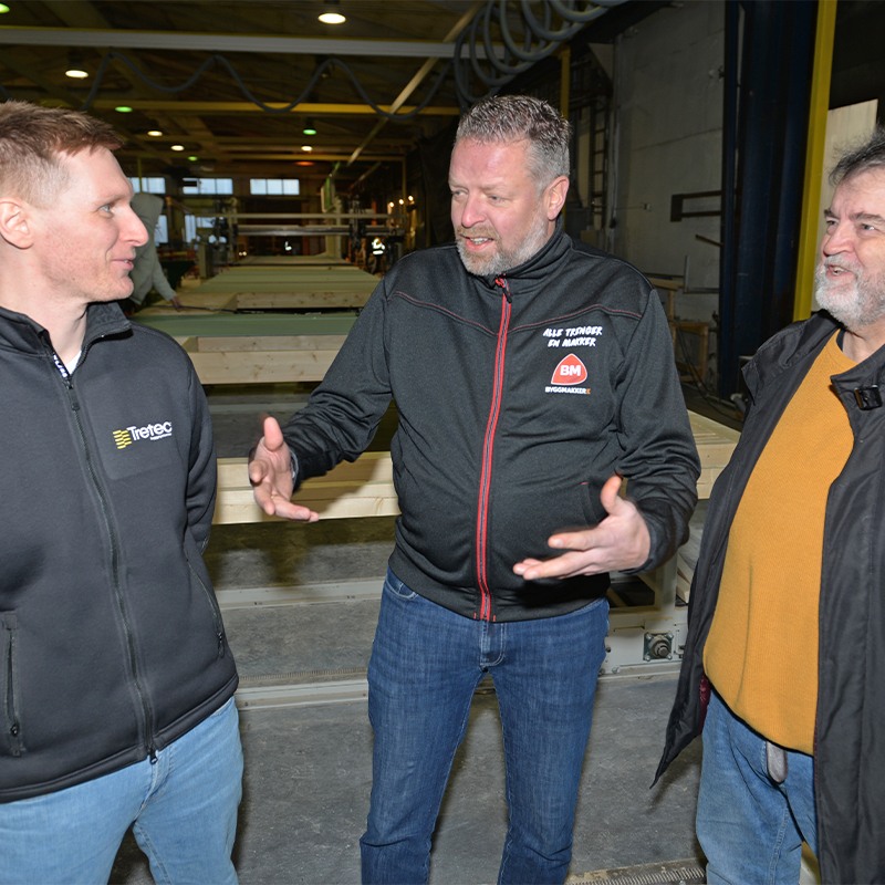 Tre menn i mørke jakker diskuterer foran lyse tremoduler på produksjonslinje i fabrikk