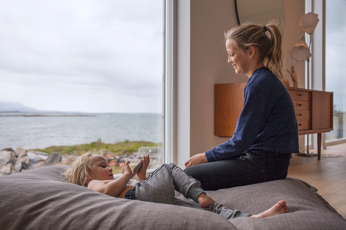 Kvinne ved stuevindu ser barn på grå saccosekk med havutsikt.