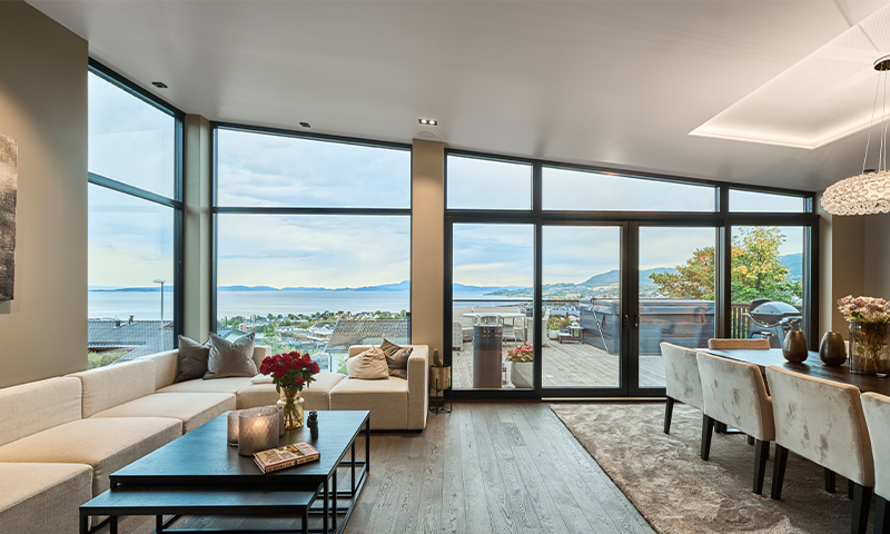 Moderne lys stue med store vinduer mot fjord, beige sofa, tremøbler og spiseplass.