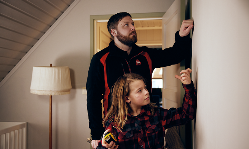 Voksen i mørk jakke og barn i rutete skjorte måler vegg med tommestokk ved lampe.