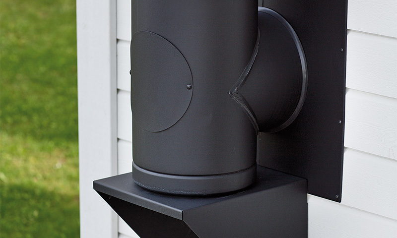 Svart metallpipe med sidegren på hvit trekledning, grønn gressplen bak