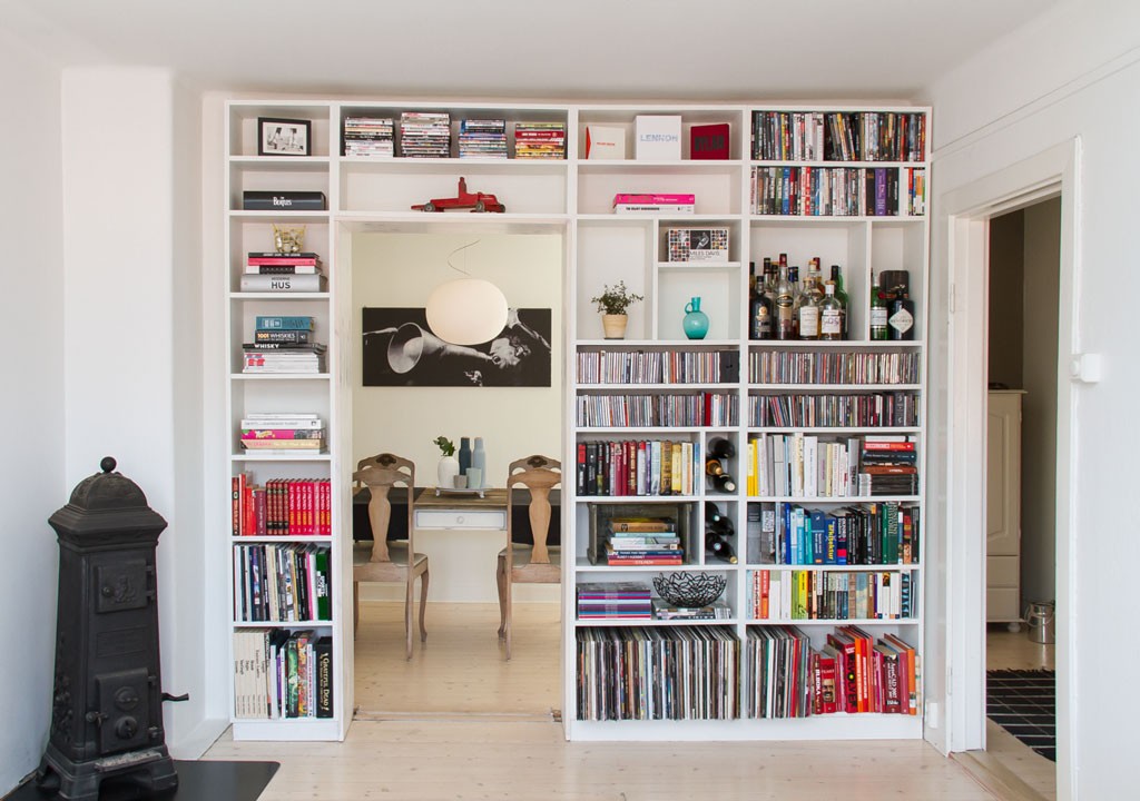 Hvit vegghylle med bøker, CD-er og flasker, åpning til rom med skrivebord, to stoler og rund lampe.