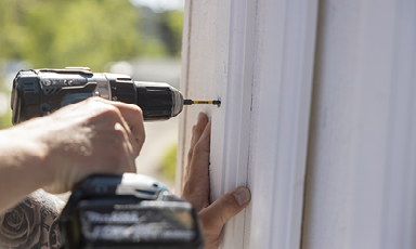 Nærbilde av en hånd som bruker svart batteridrevet drill til å skru en skrue inn i hvit trepanel.