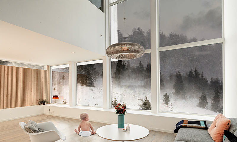 Lyst oppholdsrom med baby på tregulv, rundt hvitt bord med røde blomster, vinduer mot tåkelagt skog.
