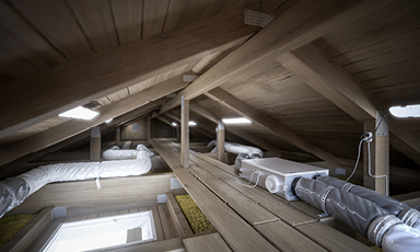 Loft med nakent treverk, synlige bjelker og isolerte grå-hvite ventilasjonsrør.