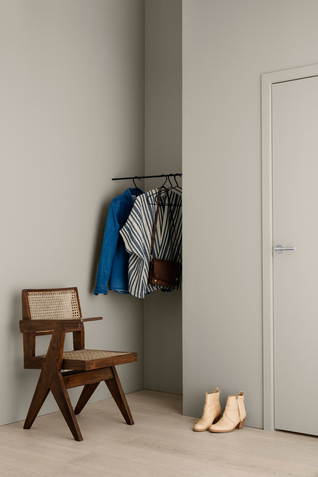 Minimalistisk ganghjørne med grå vegger, trestol, blå jakke, stripet genser, veske og beige boots.