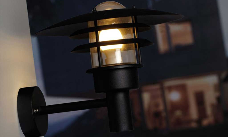 Svart utelampe i metall med varmglødende pære montert på hvit vegg ved skumring.