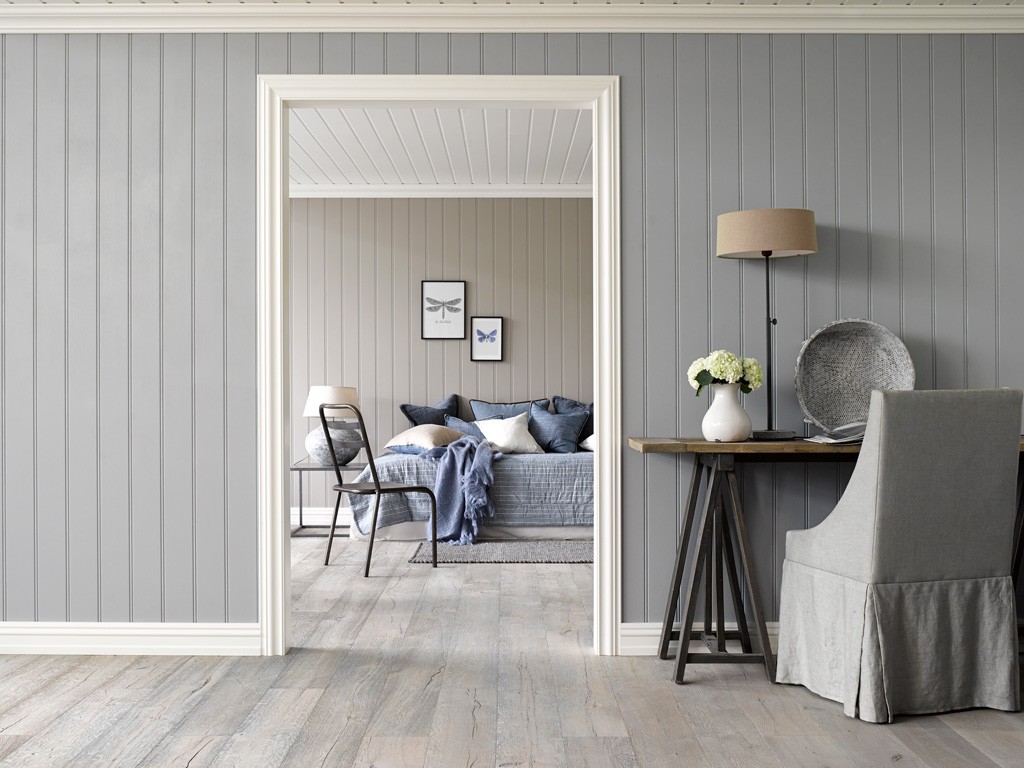 Grå stue med trebord, vase med hvite blomster og lampe. Sikt inn til soverom med blått sengeteppe.