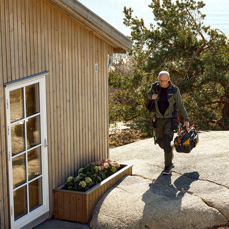 Håndverker i grønn arbeidstøy bærer verktøyveske mot lyst tømmerhus ved kysten.