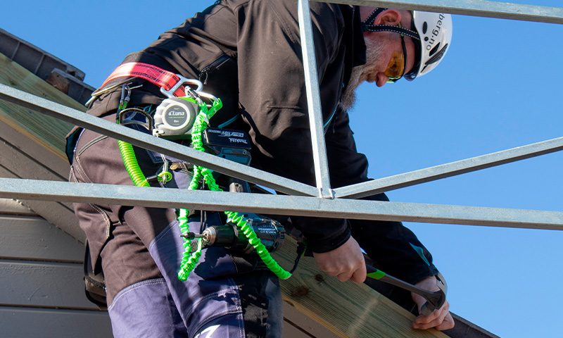 Montør i svart klær med hvit hjelm og grønt sikringsline driller metallramme mot blå himmel.
