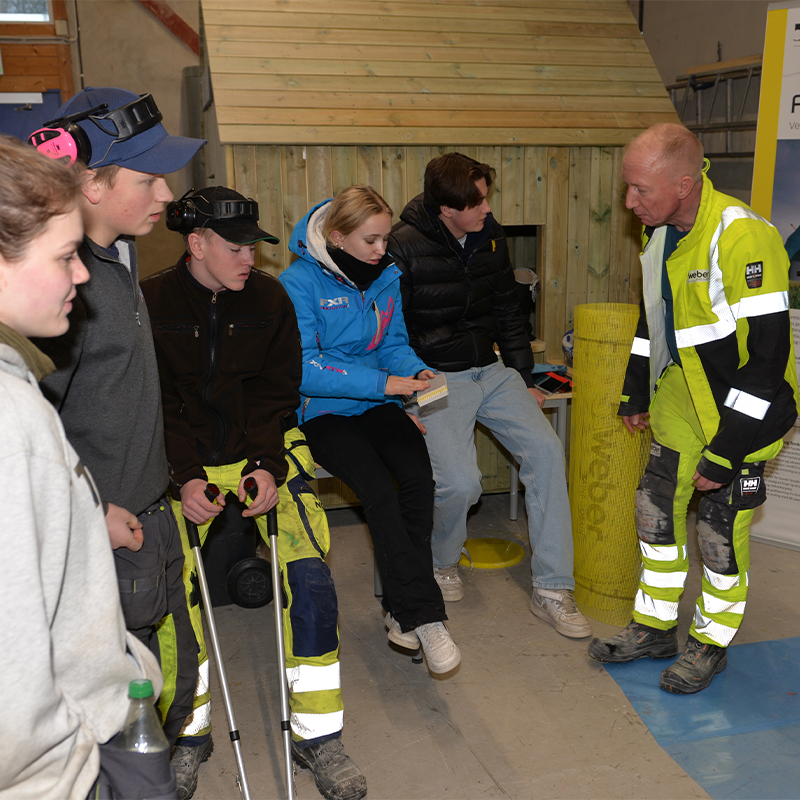 Fem personer i gule og svarte verne- og refleksklær studerer trekledning i verksted