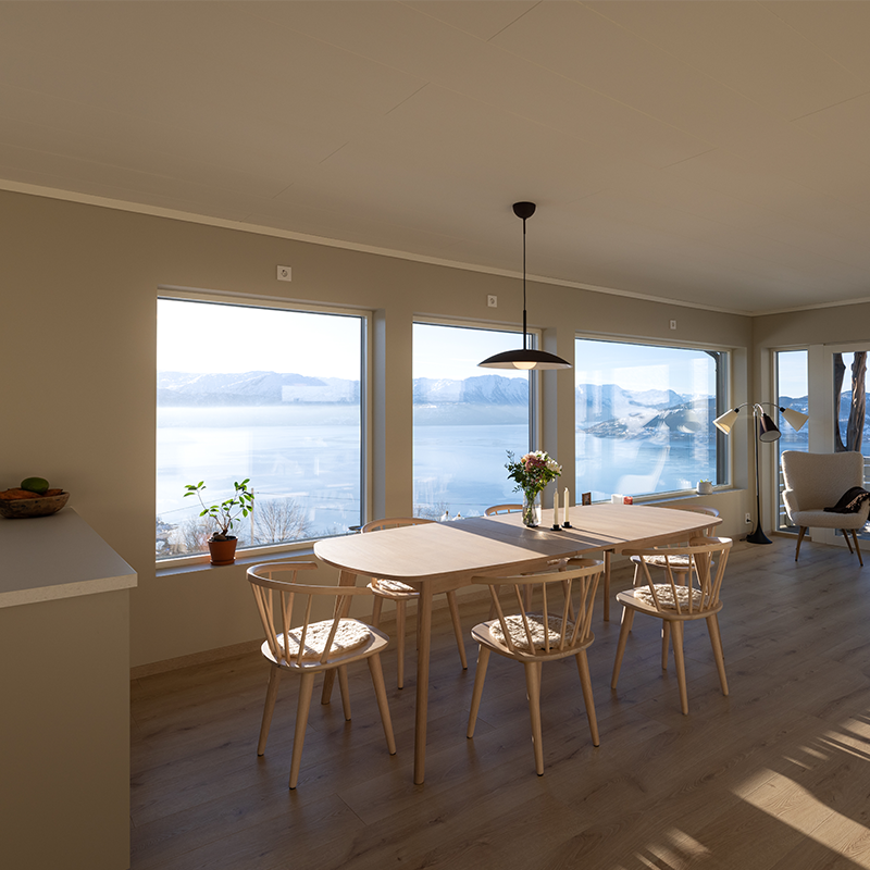 Lys spisestue med trebord og stoler, store vinduer med utsikt over fjord og fjell, og en liten blomsterbukett på bordet.