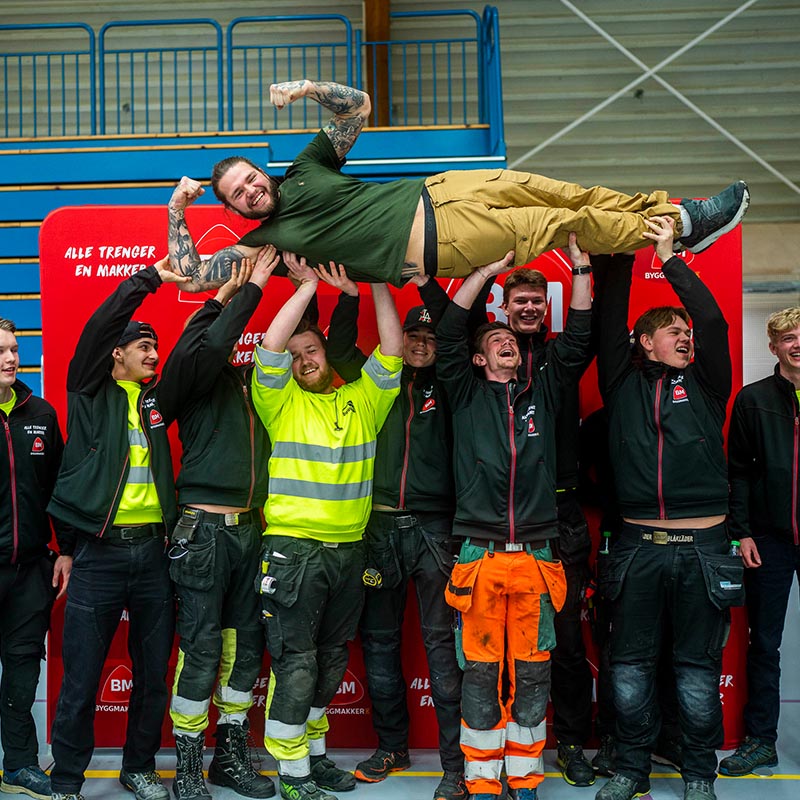 Arbeidslag i fargerike verneklær løfter kollega i grønn t-skjorte foran rødt Byggmakker-banner