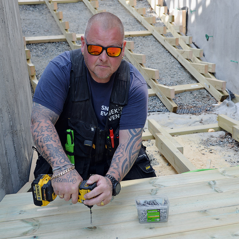 Tatovert håndverker i blå t-skjorte bruker gul drill på nytt tredekk under bygging