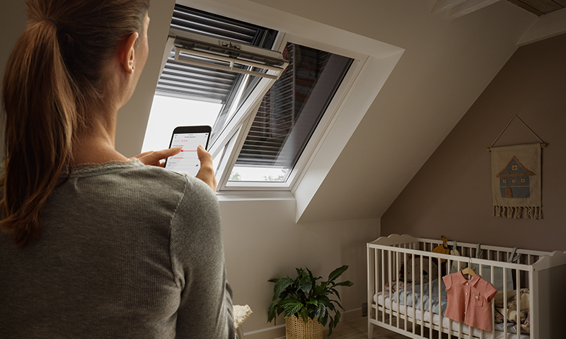 Kvinne styrer takvindu med mobil i barnerom med hvit sprinkelseng og grønn plante