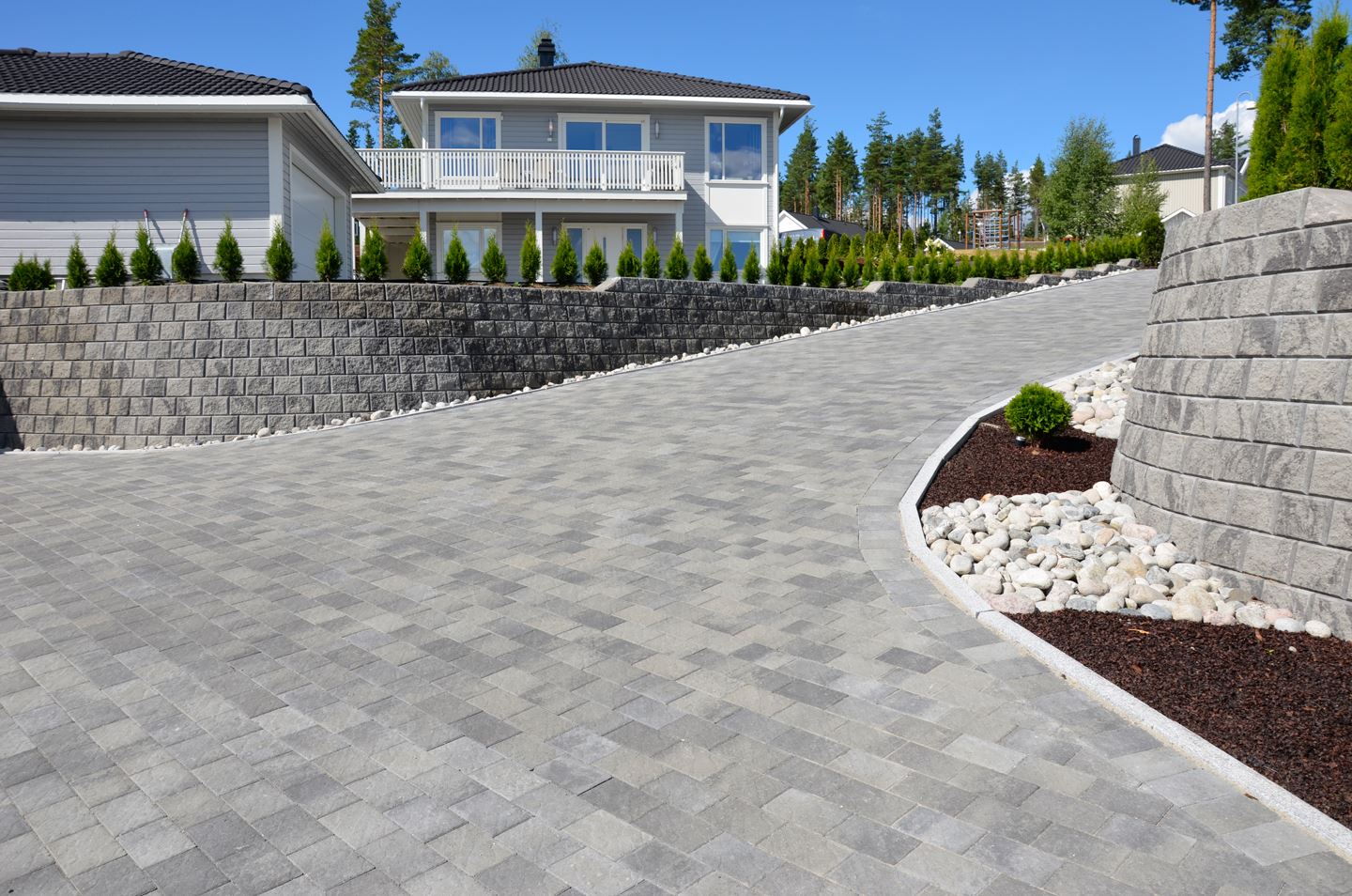 Moderne grå villa med oppkjørsel i lysegrå fliser, lav tujahekk, steinmurer og skog.