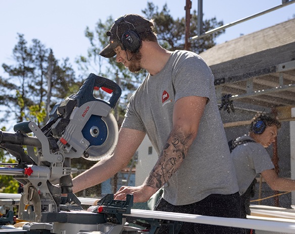 Mann i grå t-skjorte med tatovering bruker blå-hvit kapp- og gjærsag utendørs på byggeplass.