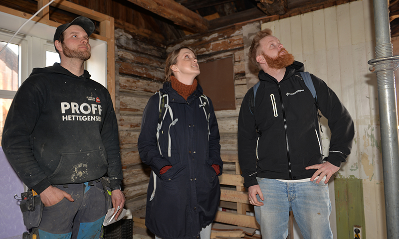 Tre personer i mørke jakker står i et uferdig, lysebrunt tømmerhus under renovering.