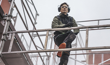 Byggearbeider i grønn jakke med hjelm og hørselsvern på metallstillas under grå himmel