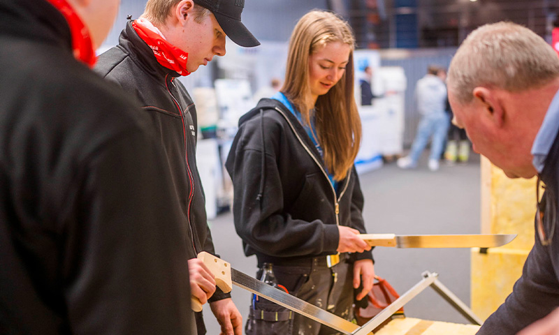 Flere personer i mørke klær tester langbladede håndsager ved en messebod.