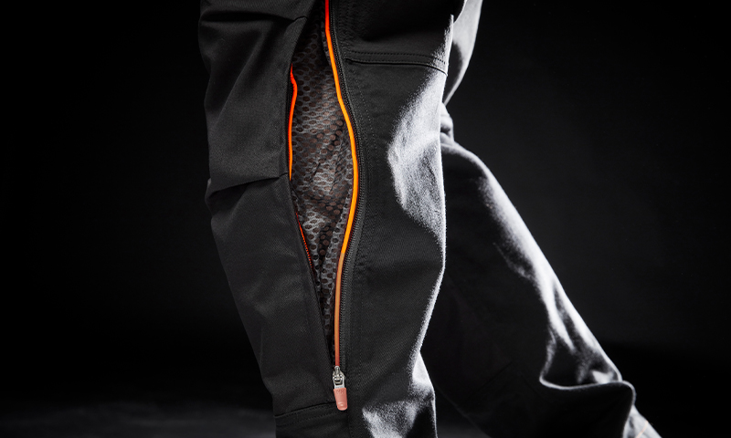 Nærbilde av svart bukseben med oransje glidelås som viser meshfôr mot svart bakgrunn.