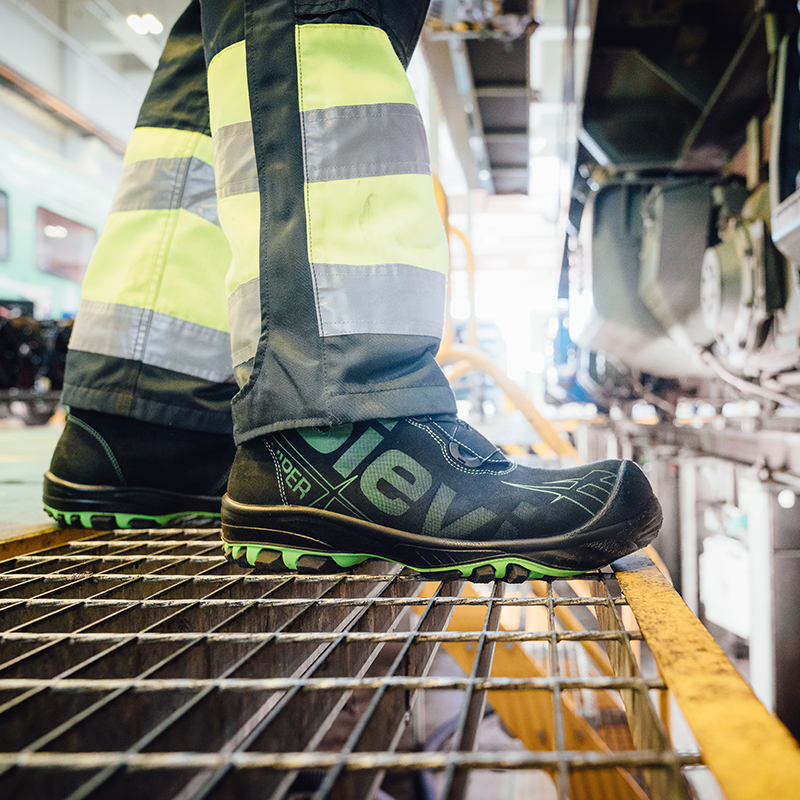Svart vernesko med grønn såle og gul refleksbukse på metallrist i industrihall.