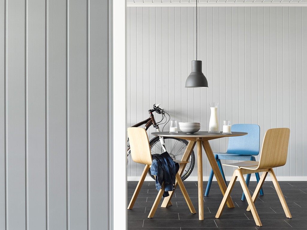 Lyst spiserom med grå vegg, trestoler (en blå), rundt bord med melk, glass og sykkel i bakgrunnen