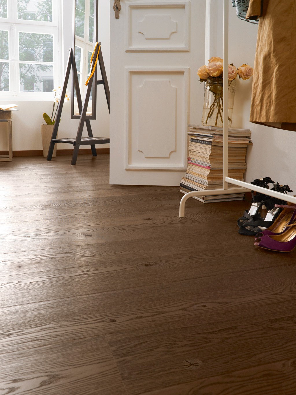 Lyst rom med mørkt tregulv, hvit dør, staffeliespeil, rosa roser på stabler magasiner og sko.