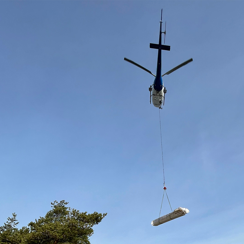 Blå-hvitt helikopter løfter innpakket byggemateriell under klar blå himmel