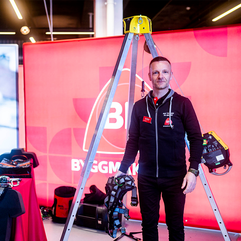 Salgsperson i svart hettegenser med sikkerhetssele foran rødt Byggmakker-banner og aluminiumstativ