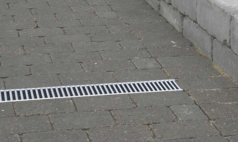 Grå belegningsstein med hvit metallrist over avløpsrenne ved murkant.