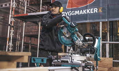 Arbeider i sort med gult hørselsvern bruker turkis kappsag foran rødt Byggmakker-banner.