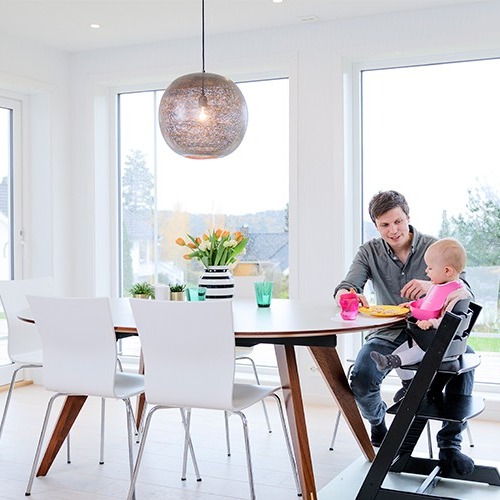 Lyst spiserom med store vinduer, hvit spisegruppe, trebord med tulipaner, mann mater baby i høy stol