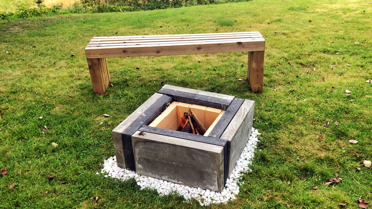 Grått, firkantet betongbålsted med hvite småstein, vedkubber og enkel trebenk på grønt gress.