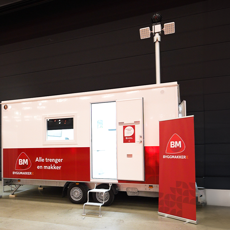 Hvit og rød Byggmakker-vogn innendørs med åpen dør, BM-banner og slogan “Alle trenger en makker”