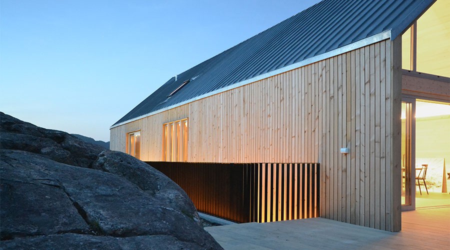 Moderne trehus med varm interiørbelysning, grått metalltak og steinutspring under blå kveldshimmel