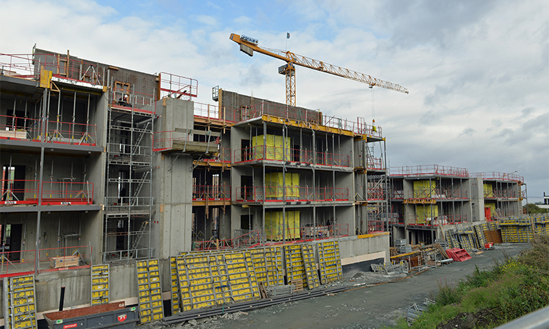 Etasjers betongbygg under oppføring, gule forskalingsplater, rødt rekkverk og kran mot skyet himmel