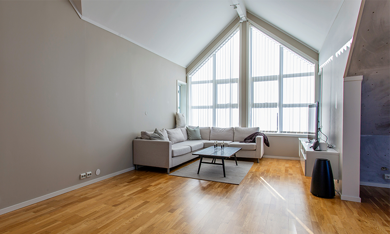 Lys stue med grå L-sofa, svart sofabord, store skråvinduer og lyst tregulv.