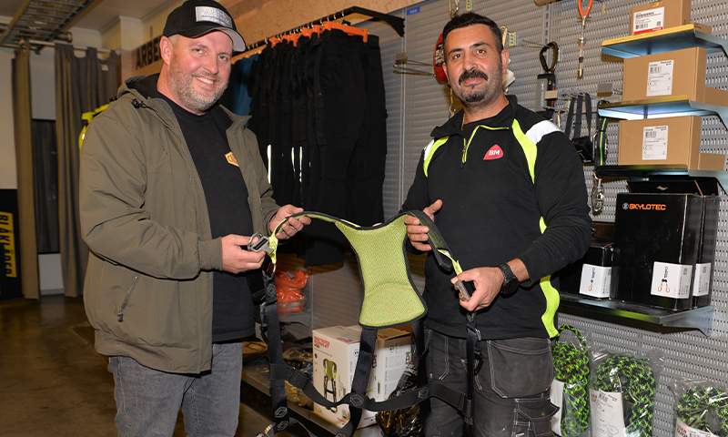 To menn i arbeidsklær i butikk, holder grønt sikkerhetssele foran hyller med utstyr.