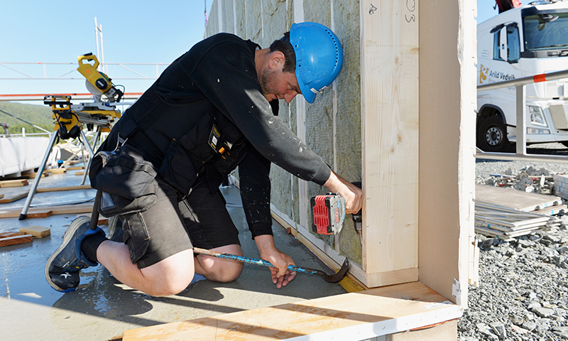 Bygningsarbeider i blå hjelm og svarte klær monterer trepanel ved byggeplass ute.