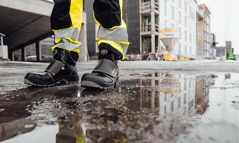 Svarte vernestøvler og gule refleksbukser på våt byggeplass med nybygg i bakgrunnen