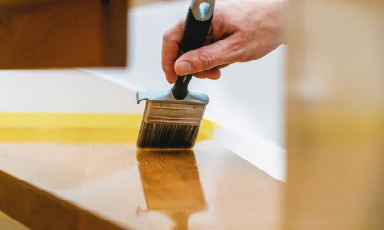 Hånd med pensel legger klar lakk på treplate, gul malertape langs kanten