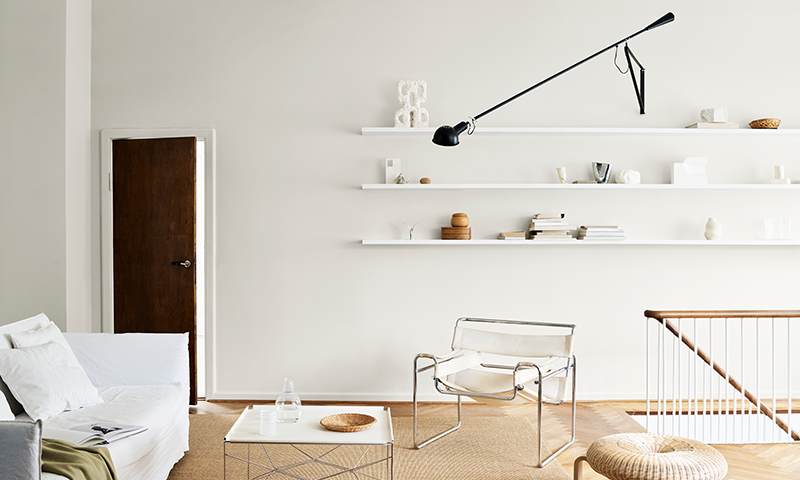 Lyst minimalistisk oppholdsrom med hvit sofa, metallstol, åpne hvite hyller og svart lampe