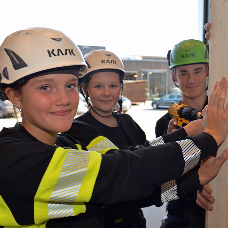 Tre personer i hvite og grønne hjelmer og gule refleksjakker fester en tømmerstokk med drill.